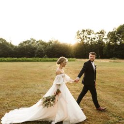 Gajówka Obręb - Para młoda, panna młoda w sukni z bufiastymi rękawami i długim trenem oraz pan młody w czarnym garniturze, trzymają się za ręce podczas spaceru po łące w promieniach zachodzącego słońca.