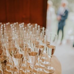 Gajówka Obręb - Rząd kieliszków do szampana, wypełnionych do połowy, ustawionych na okrągłym drewnianym stole. W tle, za rozmytą parą, ciemnoczerwona ściana.