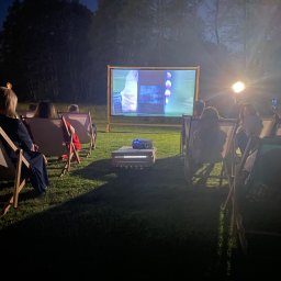 Gajówka Obręb - Kino plenerowe na trawie: widzowie siedzą w leżakach przed ekranem projekcyjnym, na którym wyświetlany jest film, oświetlonym przez reflektor, w tle drzewa i wieczorne niebo.