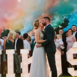 Para młoda całuje się na tle kolorowych dymów i podświetlanych liter, goście weselni w tle, oświetlenie girlandami żarówkowymi.