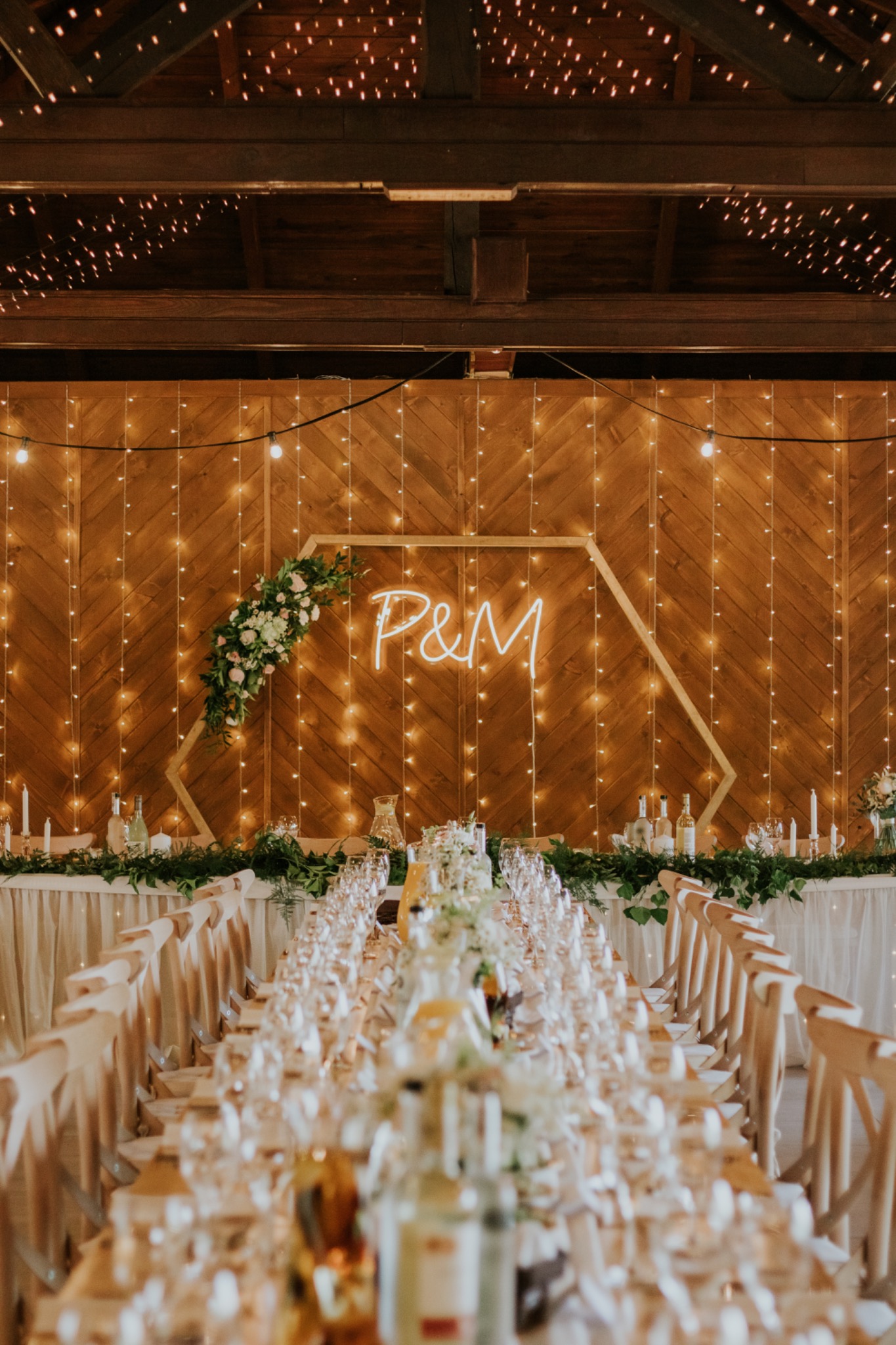 Uroczysta aranżacja stołu weselnego z długim rzędem nakryć, dekoracją z zieleni i kwiatów, drewnianym tłem z neonowym napisem 'P&M' oraz girlandami światełek.