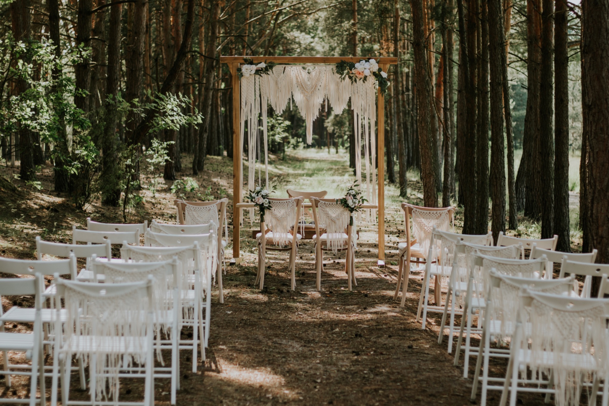 Uroczyste ustawienie krzeseł i drewnianej konstrukcji z makramą i kwiatami w leśnej scenerii, przygotowane na ceremonię ślubną.