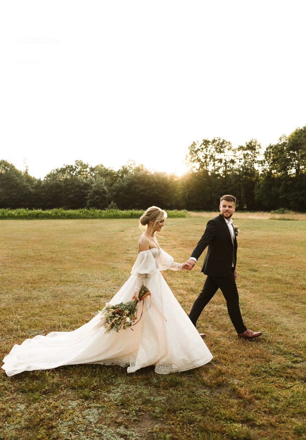 Para młoda, panna młoda w sukni z bufiastymi rękawami i długim trenem oraz pan młody w czarnym garniturze, trzymają się za ręce podczas spaceru po łące w promieniach zachodzącego słońca.