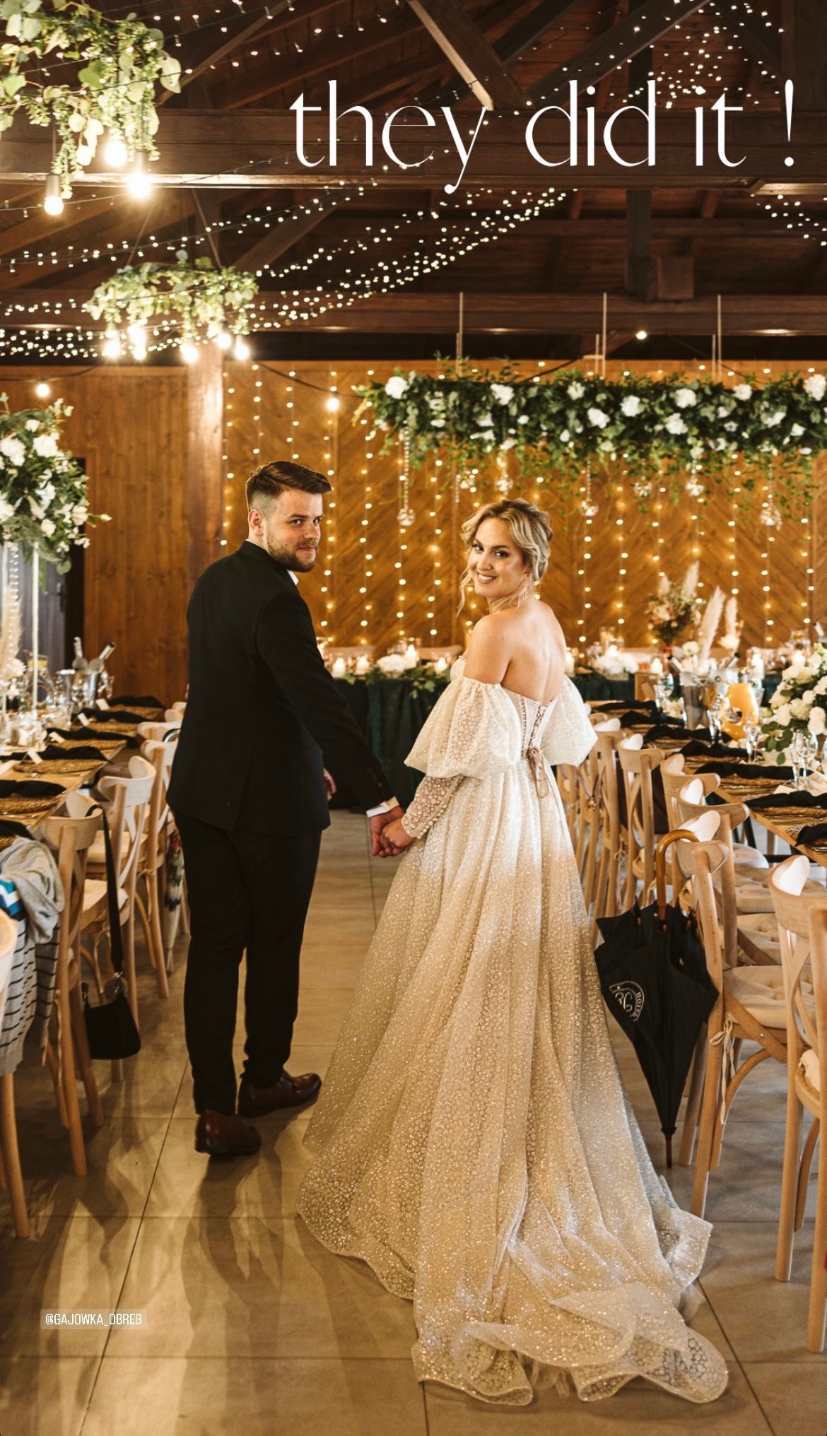 Młoda para trzymająca się za ręce, odwrócona w stronę obiektywu, na tle sali weselnej z drewnianym wystrojem, girlandami światełek i bogato zdobionymi stołami.