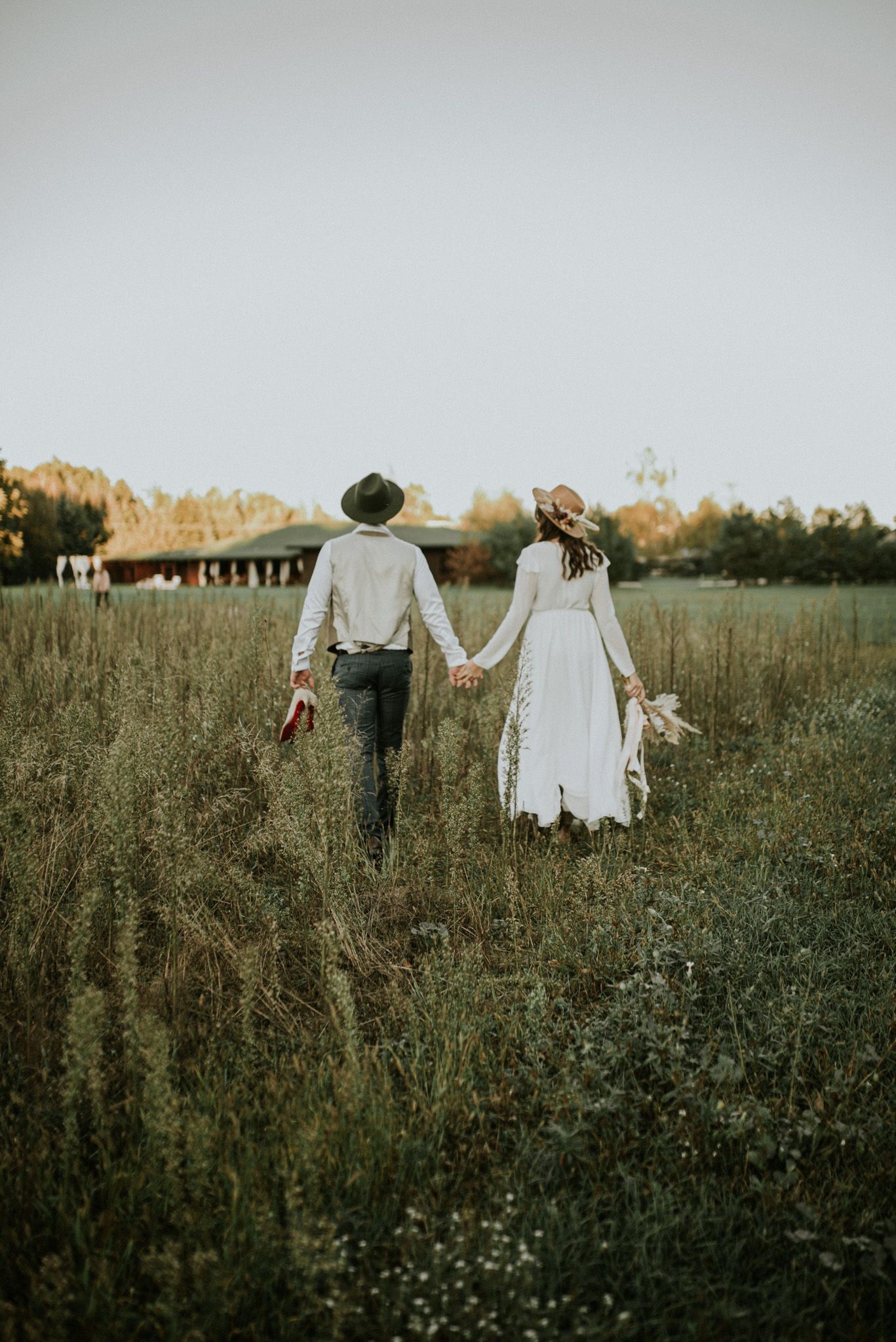 Para młoda, widziana od tyłu, idzie trzymając się za ręce przez pole z wysoką trawą w kierunku budynku z ciemnego drewna; mężczyzna trzyma czerwone buty.