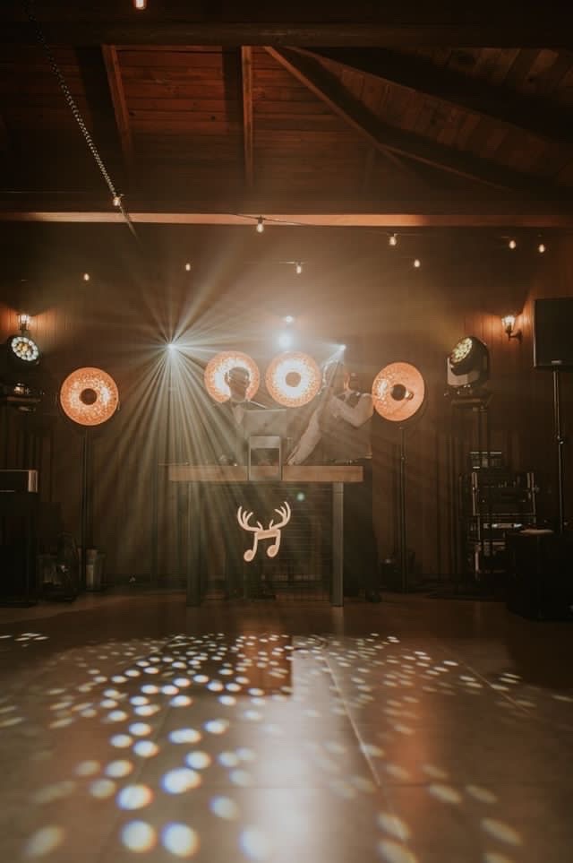 DJ-e na imprezie w rustykalnym stylu, podświetlony panel z motywem poroża i nuty, światła dyskotekowe na podłodze, oświetlenie punktowe na drewnianym suficie.