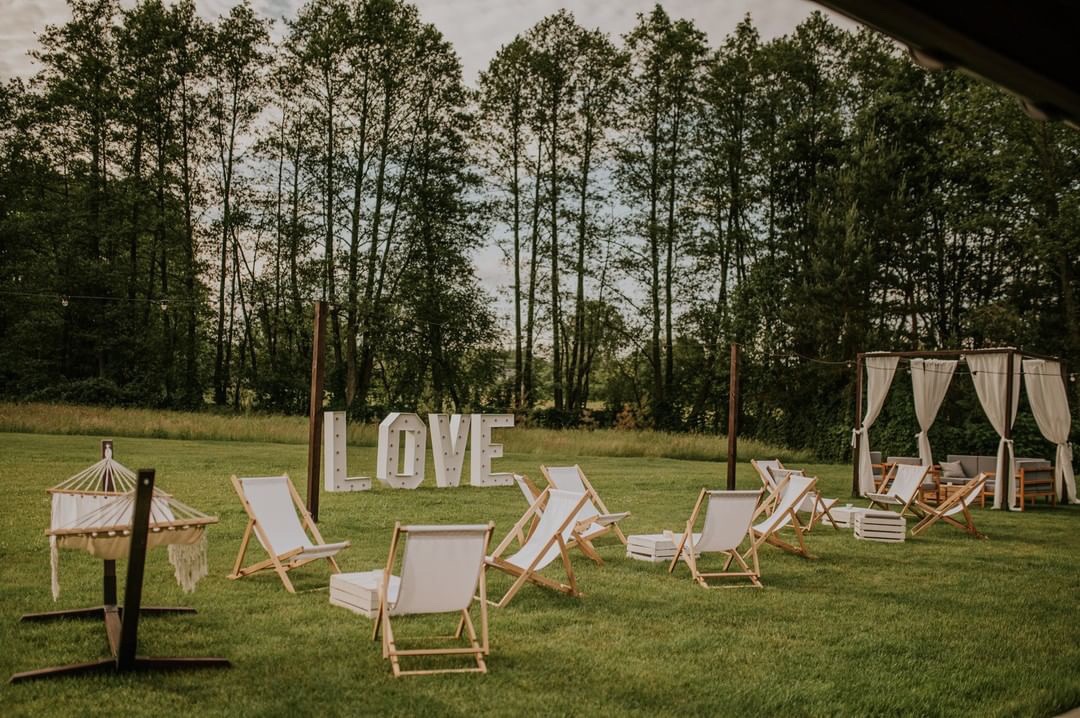 Uroczystość plenerowa z białymi leżakami, podświetlanym napisem LOVE i altaną z zasłonami na tle zielonego lasu.