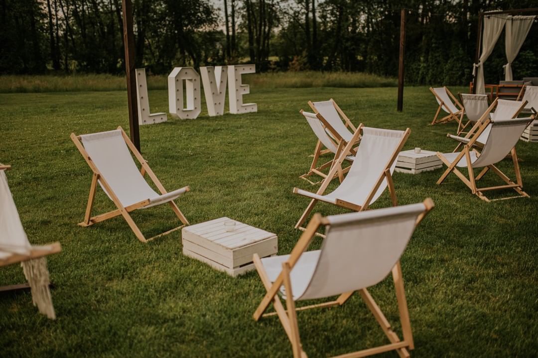 Ustawione na trawie białe leżaki z drewnianą ramą i niskie stoliki z białych skrzynek, w tle podświetlany napis LOVE, aranżacja plenerowa.