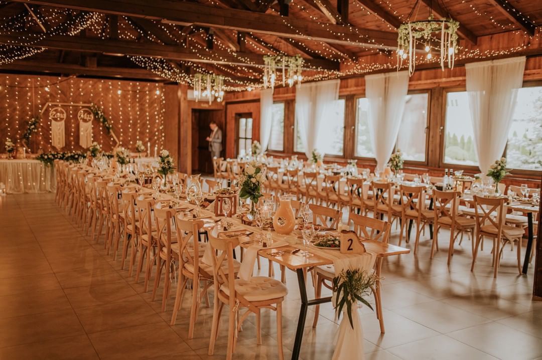 Uroczysta sala weselna z długimi stołami udekorowanymi kwiatami, numerami stołów na drewnianych podstawkach i zwisającymi girlandami z żarówek. W tle drewniane ściany i okna z białymi zasłonami.