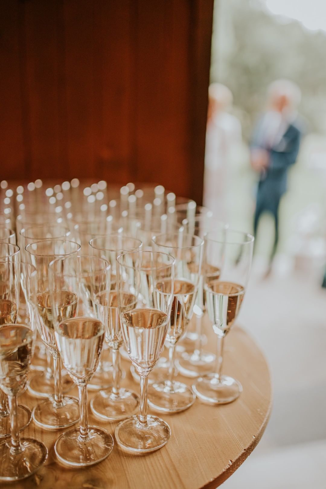 Rząd kieliszków do szampana, wypełnionych do połowy, ustawionych na okrągłym drewnianym stole. W tle, za rozmytą parą, ciemnoczerwona ściana.