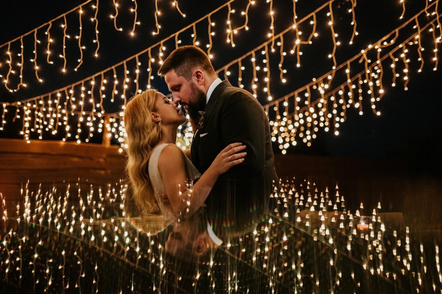 Para młoda całująca się na tle dekoracji ze światełek podczas uroczystości weselnej.