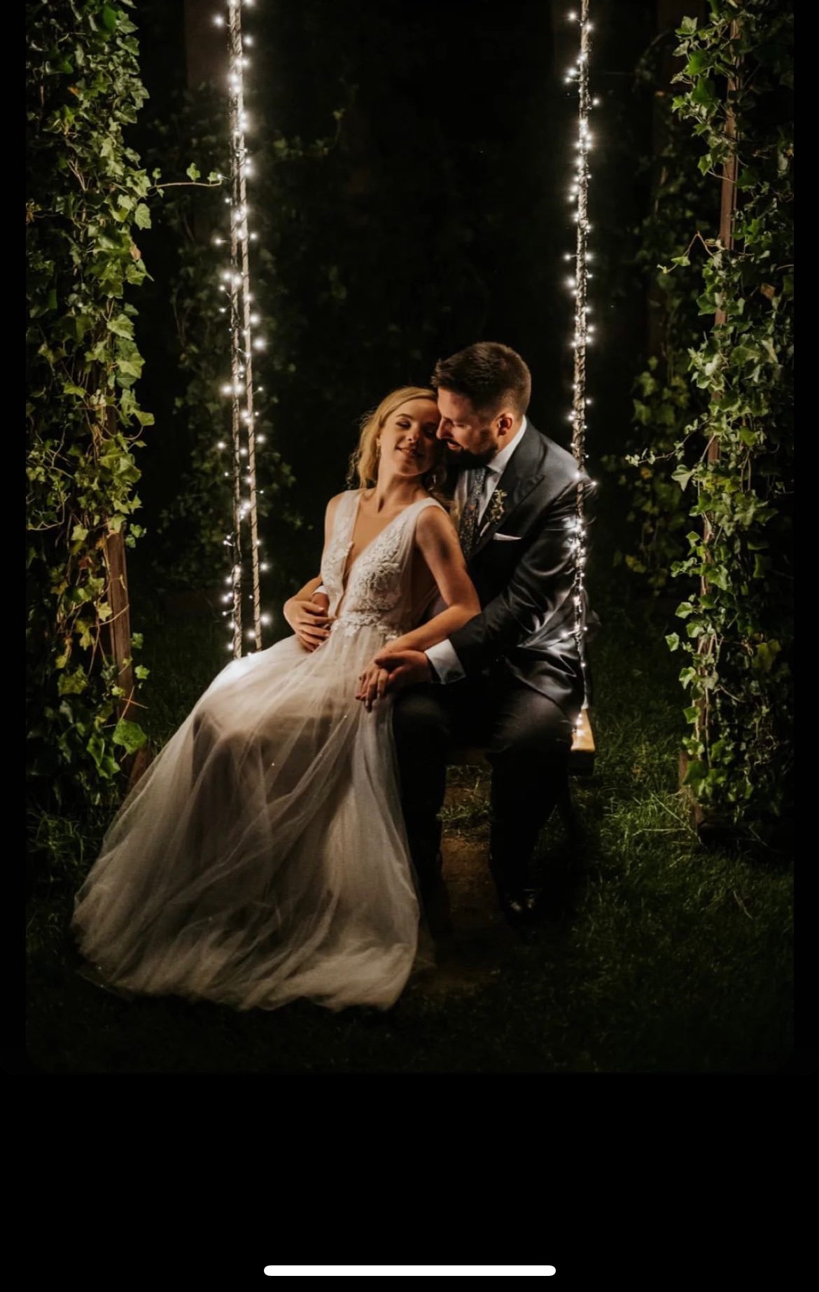 Para młoda siedząca na huśtawce ozdobionej światełkami w ogrodzie, panna młoda w koronkowej sukni z głębokim dekoltem, pan młody w ciemnym garniturze, romantyczna atmosfera.