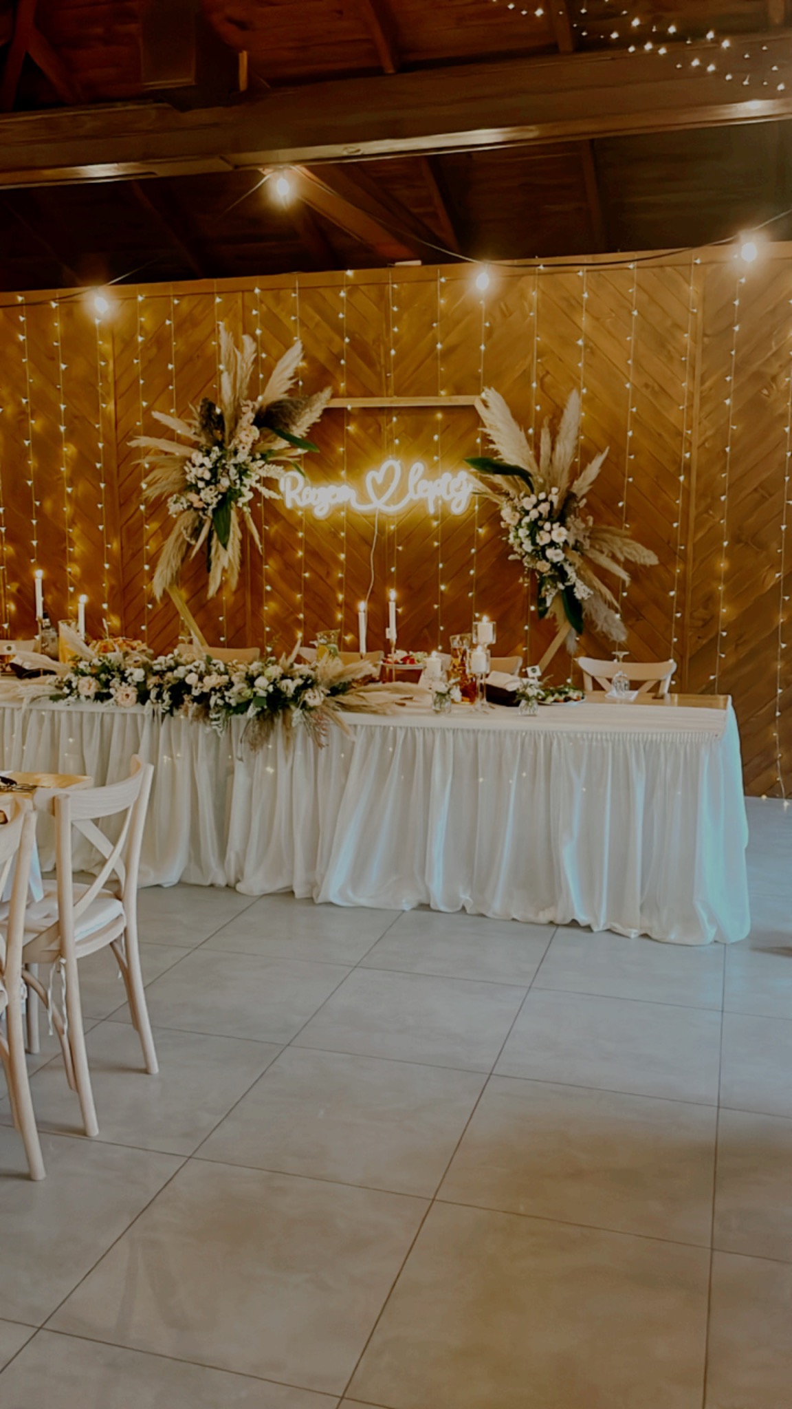 Uroczysta dekoracja stołu weselnego z białym obrusem, świeżymi kwiatami i pampasową trawą, podświetlona girlandą z żarówek i neonowym napisem 'Razem Lepiej' w drewnianej ramie o heksagonalnym...