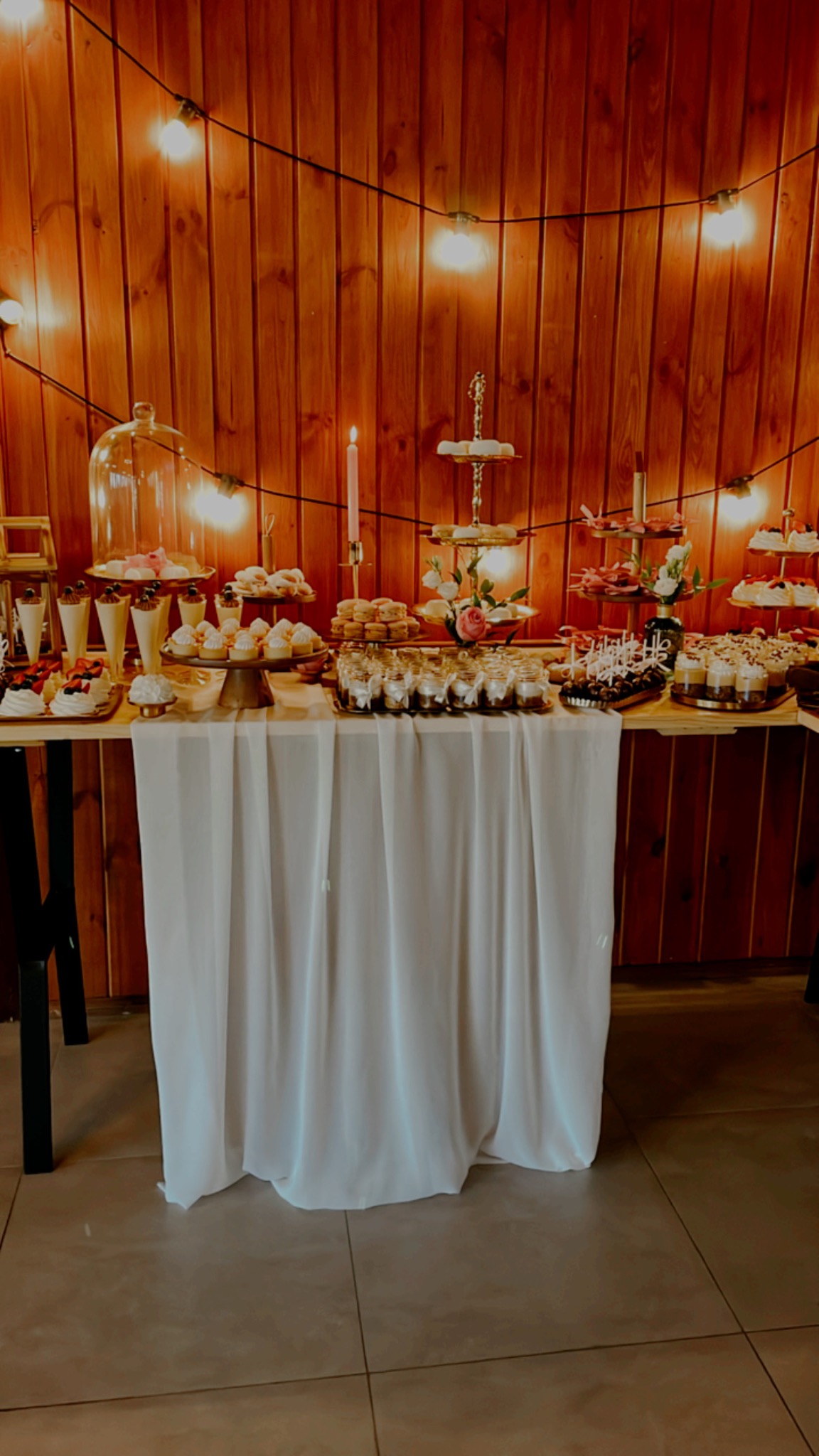 Stół z białym obrusem uginający się pod ciężarem słodkich przekąsek: babeczek, deserów w słoiczkach, mini bez i donatów, oświetlony girlandą żarówek na tle drewnianej ściany.
