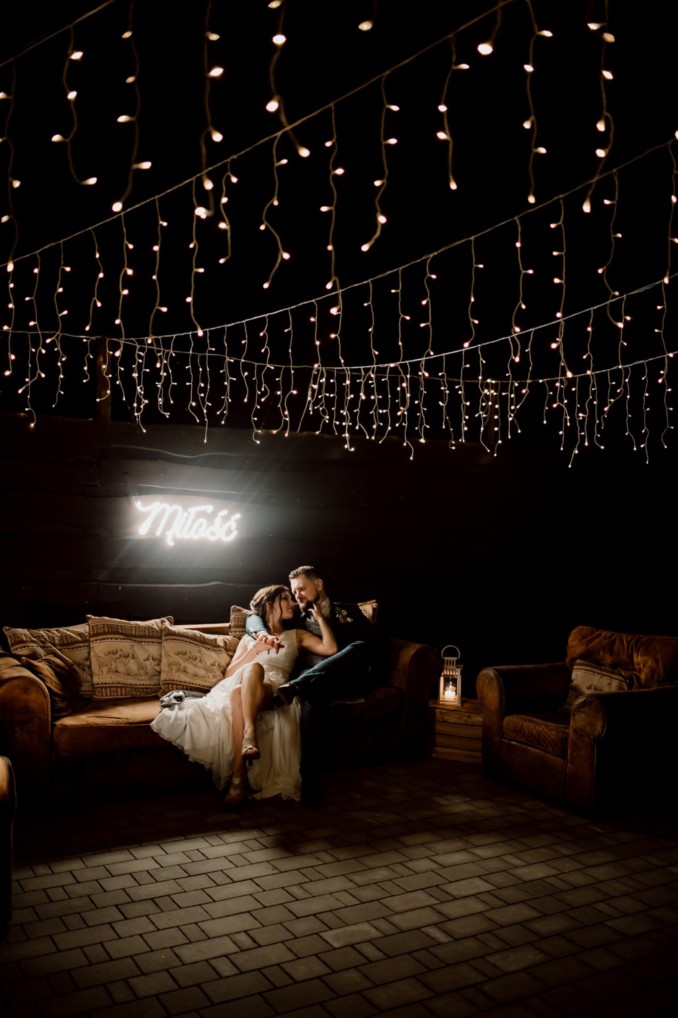 Para młoda siedzi na brązowej sofie pod girlandą światełek i neonowym napisem 'Miłość' podczas wieczornej uroczystości, obok stoi lampion ze świecą.