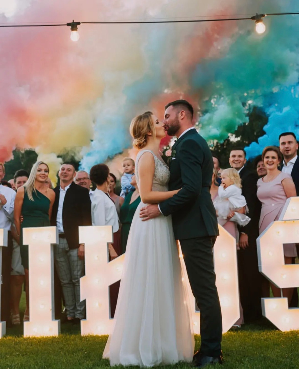 Para młoda całuje się na tle kolorowych dymów i podświetlanych liter, goście weselni w tle, oświetlenie girlandami żarówkowymi.