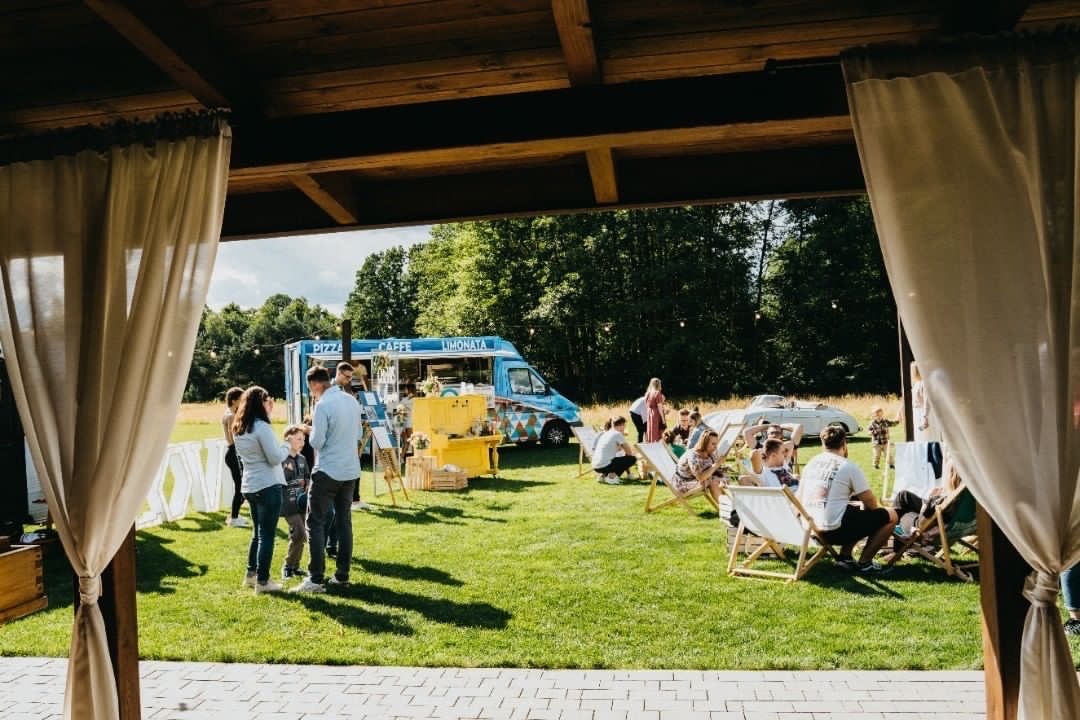 Plenerowa impreza z food truckiem serwującym pizzę, kawę i lemoniadę, goście siedzą na leżakach, widok z zadaszonej przestrzeni z białymi zasłonami.
