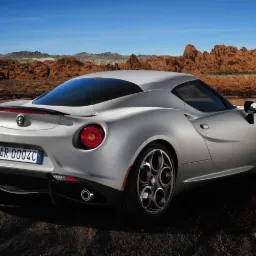 Srebrny samochód sportowy Alfa Romeo 4C Coupe z charakterystycznymi felgami, sfotografowany z tyłu na tle pustynnego krajobrazu z czerwonymi formacjami skalnymi i błękitnym niebem.