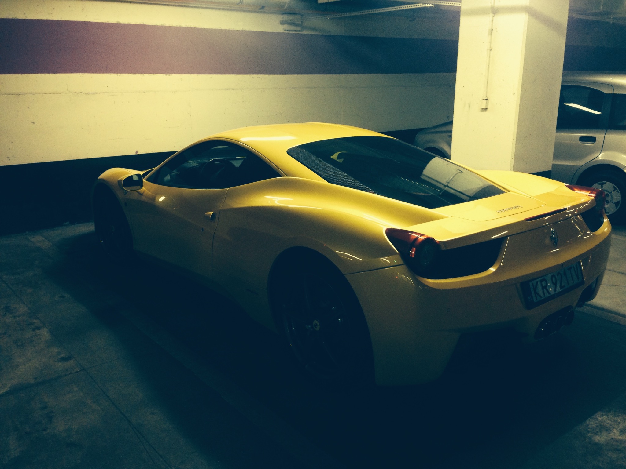 Żółty samochód sportowy marki Ferrari, zaparkowany w garażu podziemnym, widok z tyłu, z widoczną tablicą rejestracyjną i elementami nadwozia.