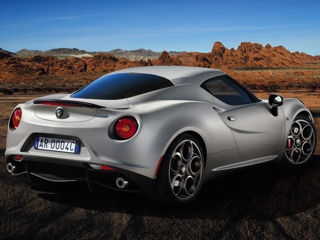 Srebrny samochód sportowy Alfa Romeo 4C Coupe z charakterystycznymi felgami, sfotografowany z tyłu na tle pustynnego krajobrazu z czerwonymi formacjami skalnymi i błękitnym niebem.