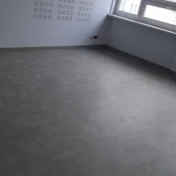 Szara, gładka posadzka jastrychowa w jasnym pomieszczeniu z dużym oknem i perforowaną ścianą akustyczną.