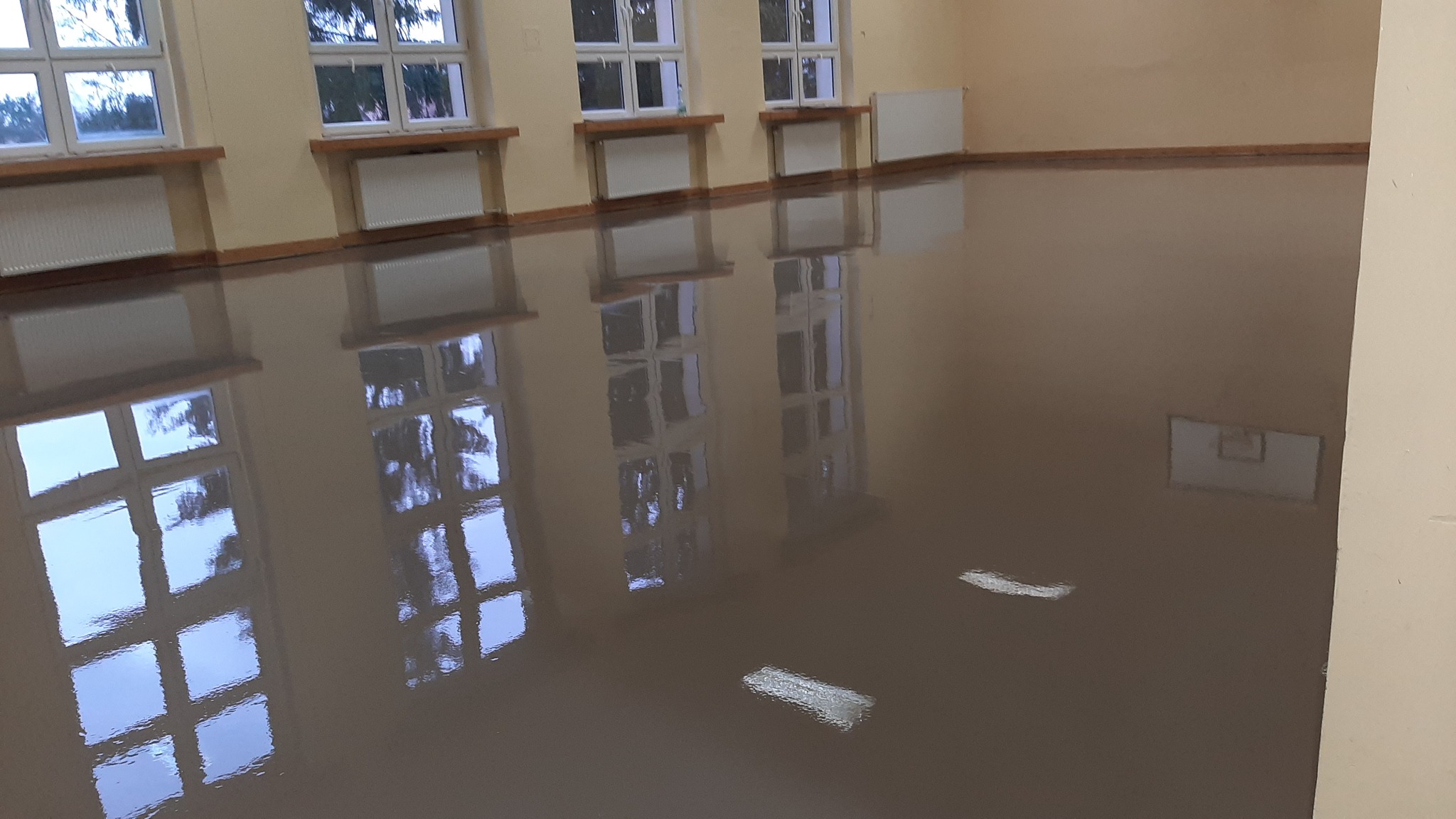 Błyszcząca, gładka posadzka w jasnym pomieszczeniu z oknami, odbijająca światło i widok z zewnątrz. Widoczne grzejniki pod oknami i listwy przypodłogowe.