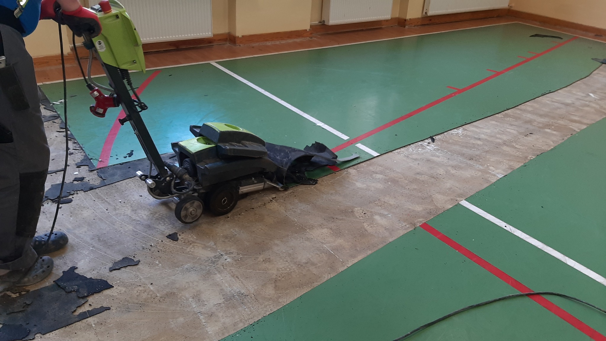 Usuwanie starej, zielonej wykładziny z podłogi hali sportowej za pomocą specjalistycznej maszyny z zielonym korpusem; widoczne fragmenty oderwanej wykładziny i ślady kleju.