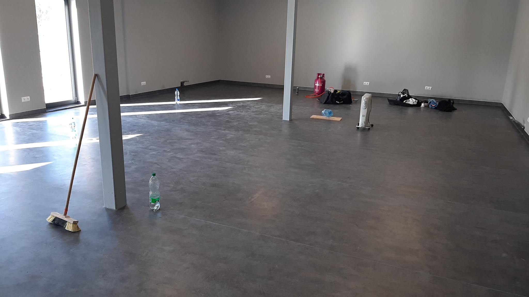 Surowa, szara posadzka w przestronnym pomieszczeniu z widocznymi śladami prac wykończeniowych: butelki z wodą, miotła oparta o filar, torby z narzędziami i nagrzewnica.