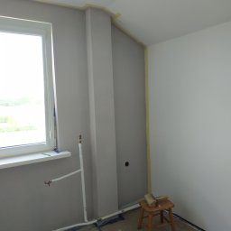 Silver Hands - Pokój w trakcie odświeżania: pomalowana na szaro ściana z widocznymi rurami, zabezpieczone taśmą malarską krawędzie i podłoga, mały drewniany stołek z pędzlem na siedzisku.