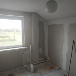 Silver Hands - Pokój w trakcie szpachlowania ścian, widoczne ślady po szpachli, okno z widokiem na zewnątrz, wisząca lampa, rury instalacji wodnej, szlifierka do gładzi i akcesoria malarskie na podłodze.