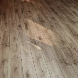 Ukończona podłoga z paneli laminowanych o wzorze jasnego dębu, ułożona wzdłuż, z widocznym fragmentem ściany z tapetą w liście w tle, oświetlona naturalnym światłem tworzącym smugę na powierzchni.