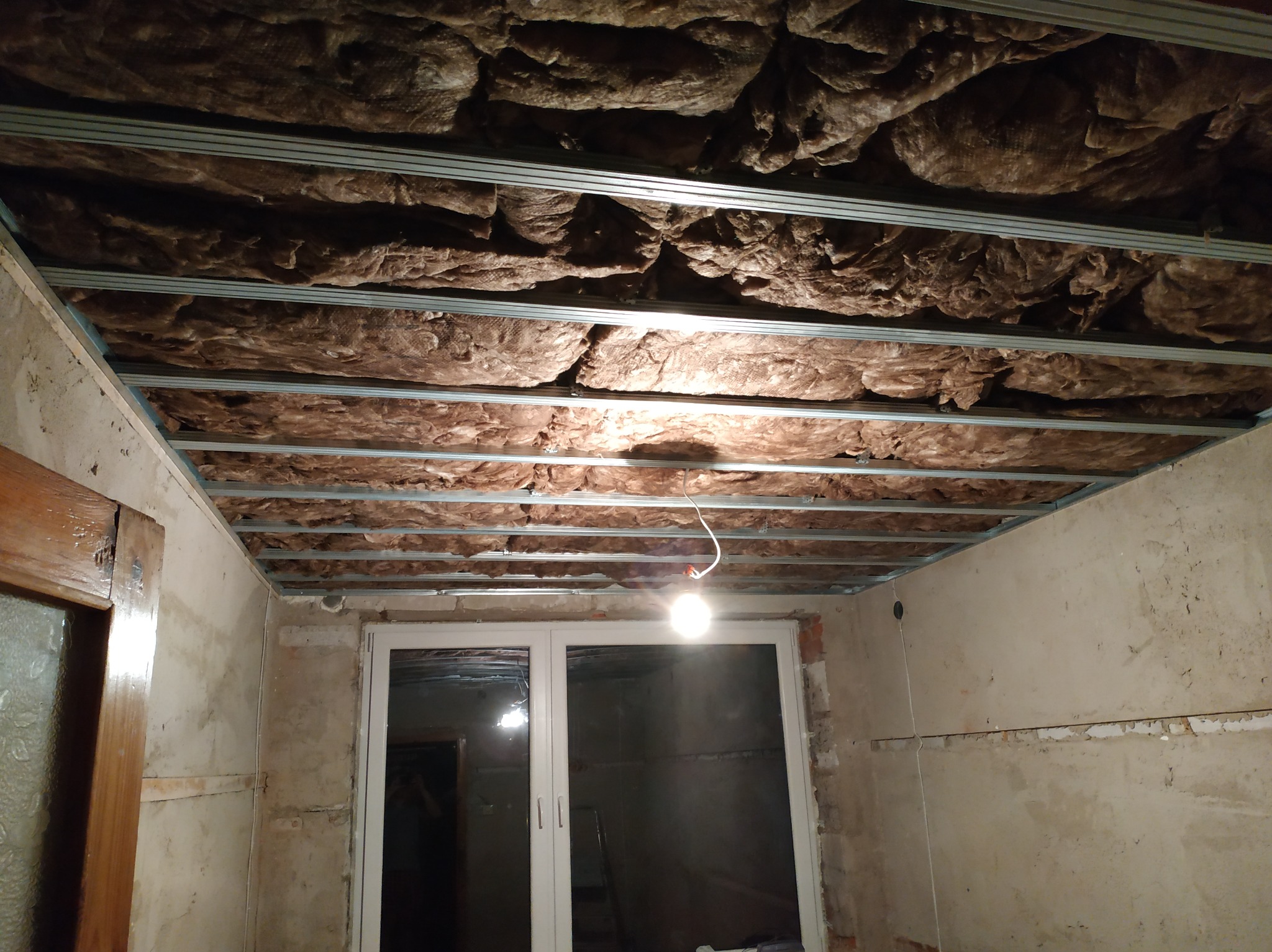 Montaż izolacji z wełny mineralnej pomiędzy metalowymi profilami podwieszanego sufitu w trakcie prac wykończeniowych, widoczna surowa ściana i okno.