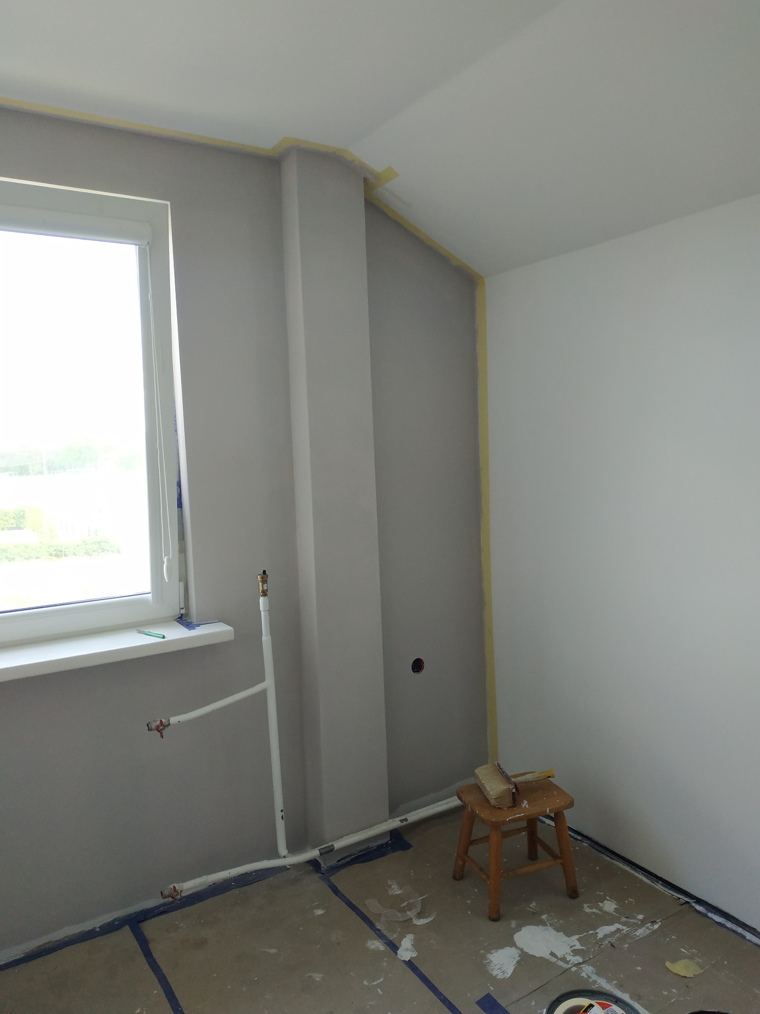 Pokój w trakcie odświeżania: pomalowana na szaro ściana z widocznymi rurami, zabezpieczone taśmą malarską krawędzie i podłoga, mały drewniany stołek z pędzlem na siedzisku.