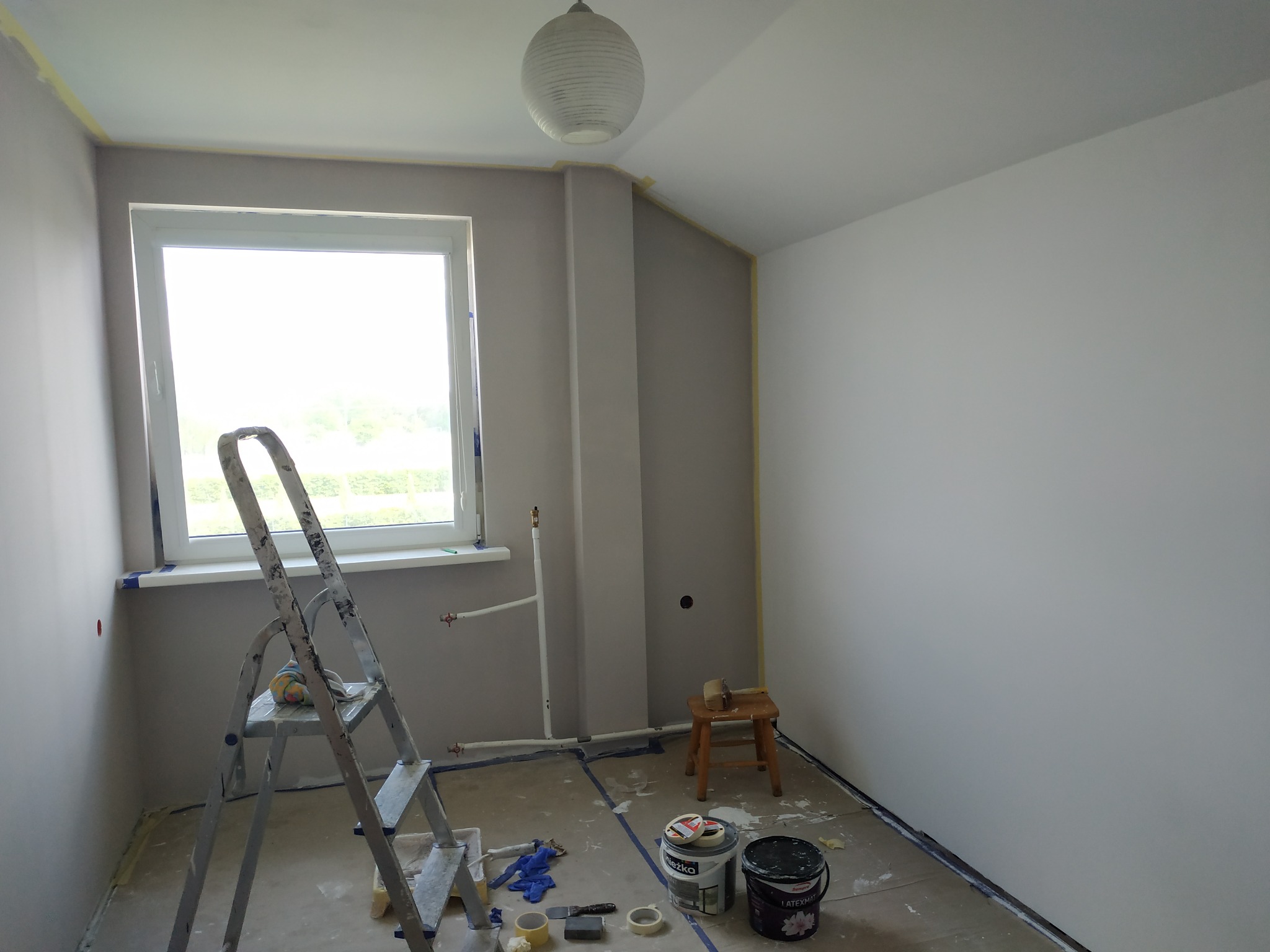 Pokój w trakcie malowania: ściany częściowo pomalowane na szaro i biało, zabezpieczone taśmą malarską, drabina, pędzle, wałki i dwa wiadra z farbą na podłodze.