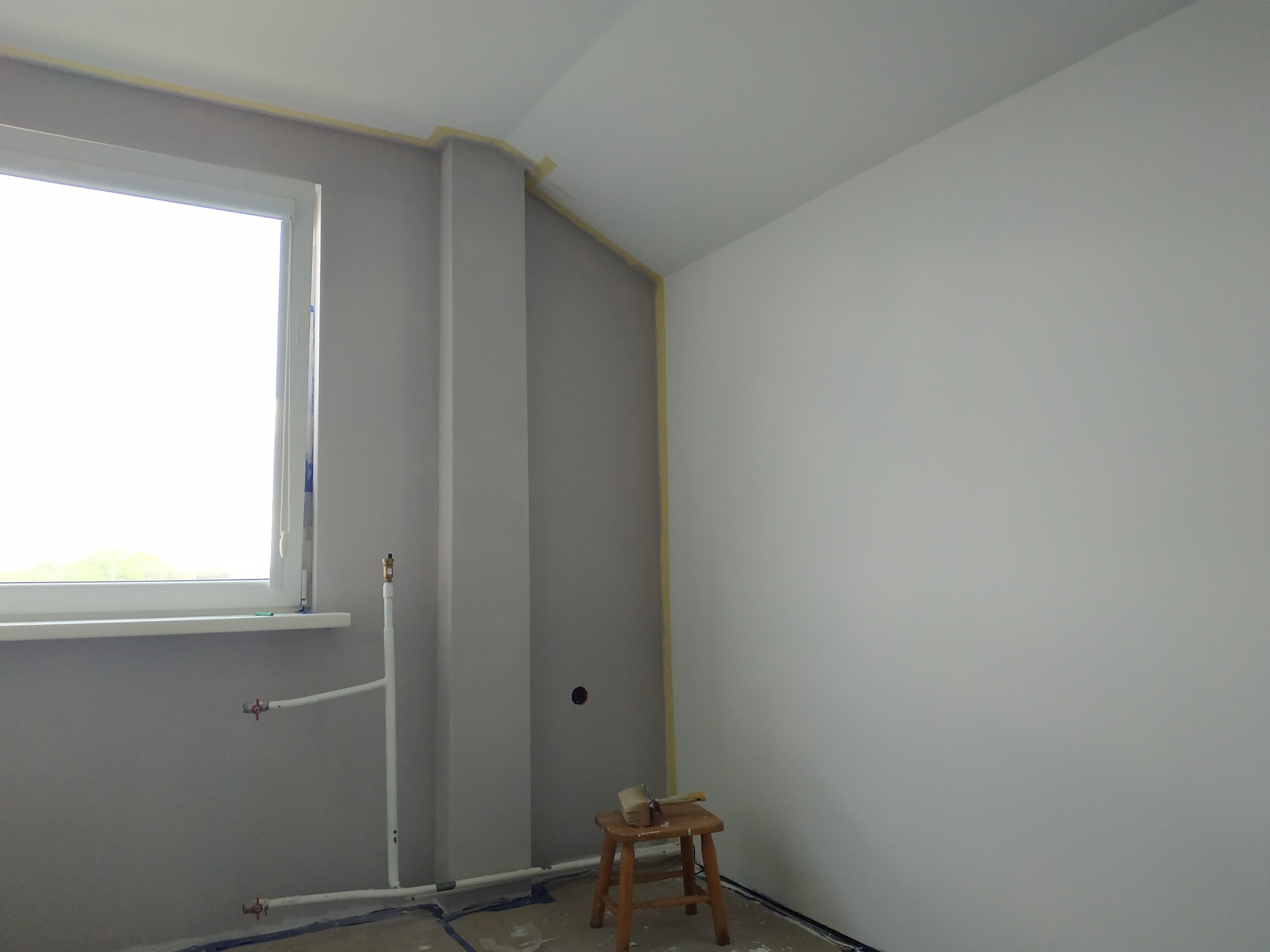 Pokój w trakcie malowania w Łodzi: ściany częściowo pomalowane na szaro, zabezpieczone taśmą malarską przy suficie, widoczne rury grzewcze przy oknie i stołek z pędzlem.