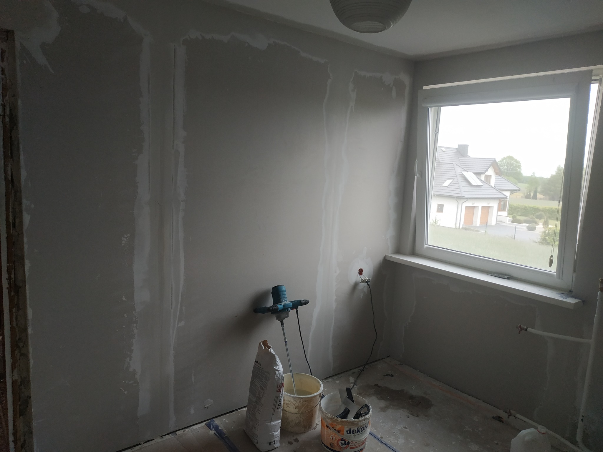Pokój w trakcie szpachlowania ścian, widoczne ślady po łatach gipsowych, wiadro z zaprawą, mieszadło elektryczne i worek gipsu.
