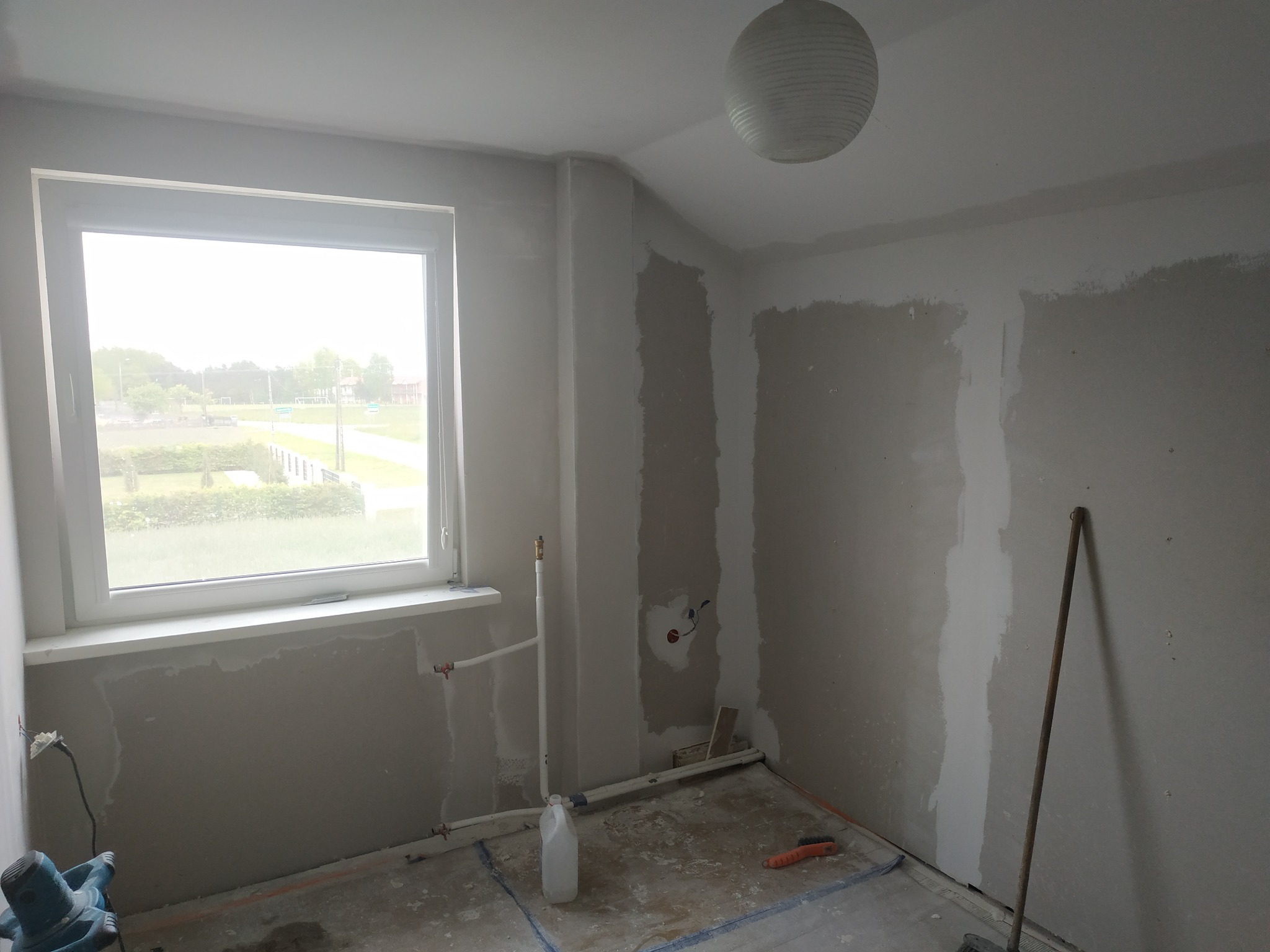 Pokój w trakcie szpachlowania ścian, widoczne ślady po szpachli, okno z widokiem na zewnątrz, wisząca lampa, rury instalacji wodnej, szlifierka do gładzi i akcesoria malarskie na podłodze.