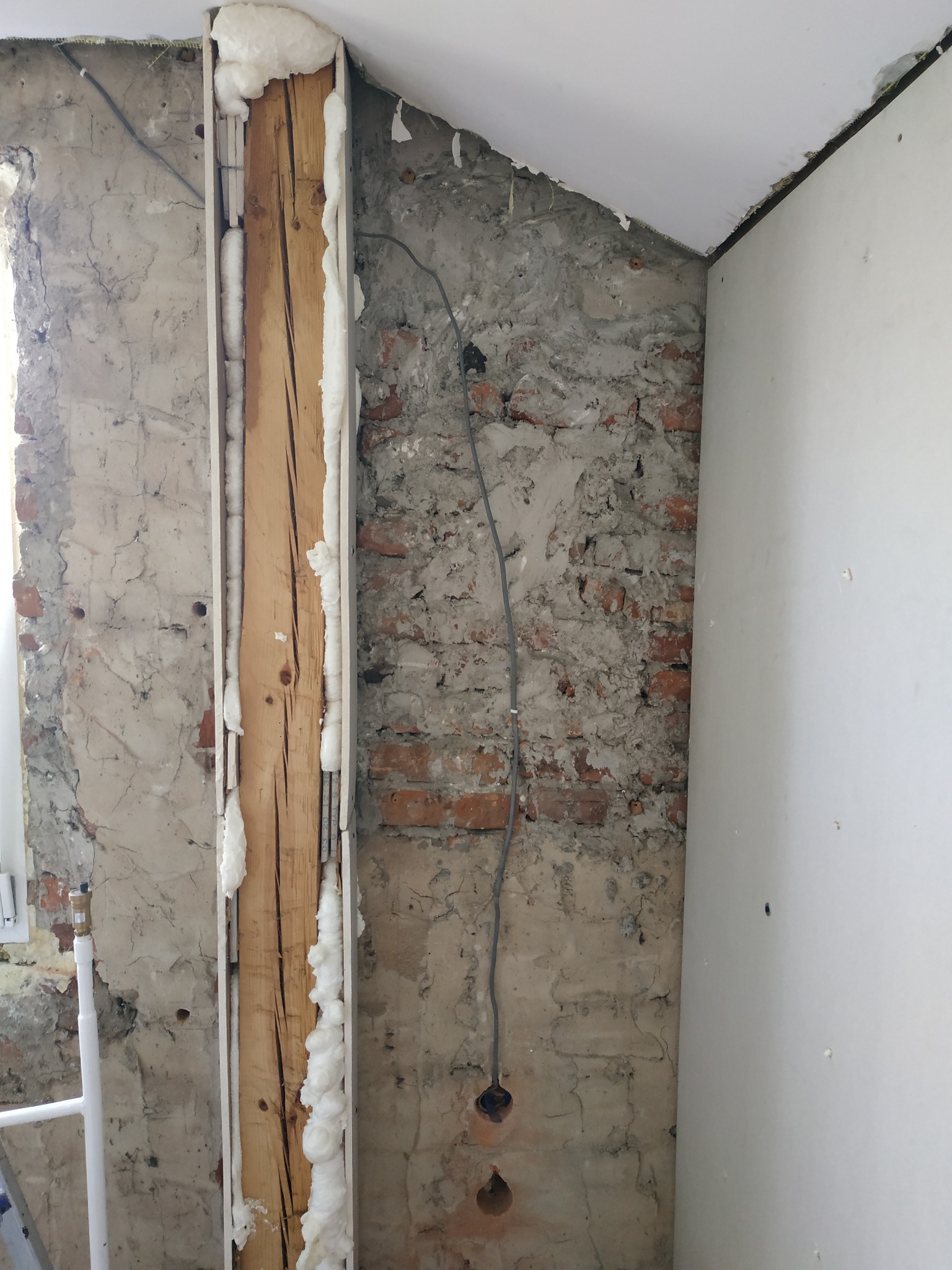 Fragment ściany w trakcie prac wykończeniowych: odsłonięta cegła, instalacja elektryczna z otworem, drewniana belka z pianką montażową oraz fragment płyty gipsowej, ukazujący etap przygotowania...