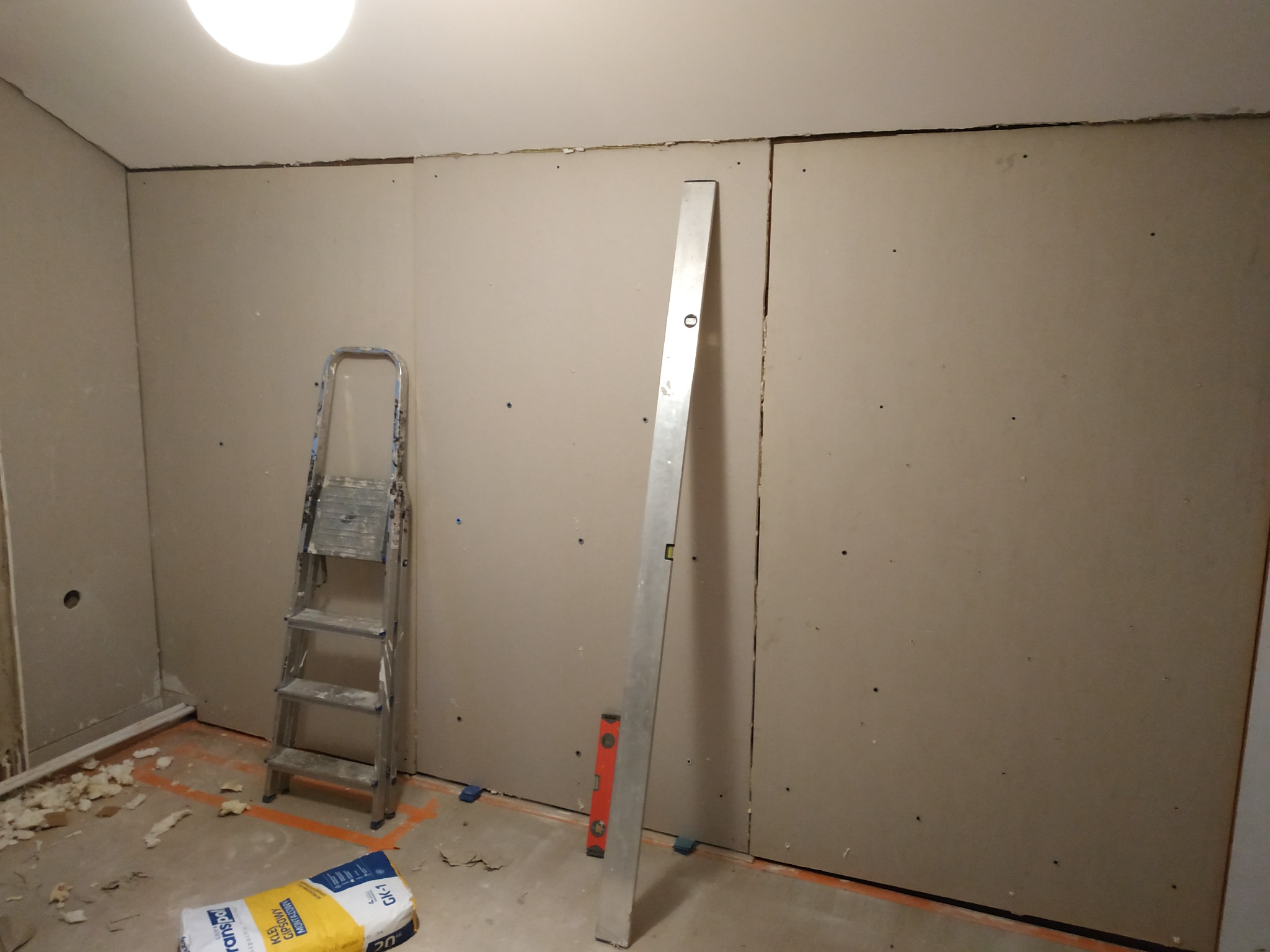 Montaż płyt gipsowo-kartonowych na ścianie z widoczną drabinką, poziomicą i workiem kleju gipsowego.