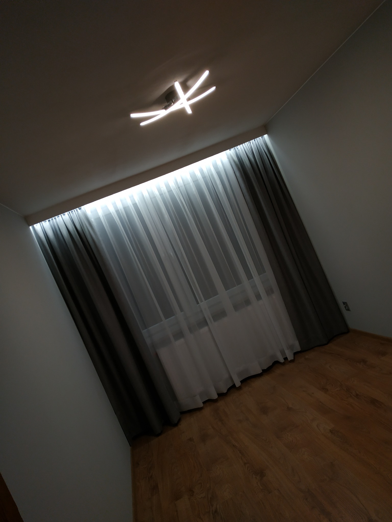 Nowoczesne oświetlenie sufitowe w kształcie krzyża z czterema ramionami, widoczne na tle białego sufitu w pokoju z drewnianą podłogą i oknem zasłoniętym szarymi i białymi zasłonami.