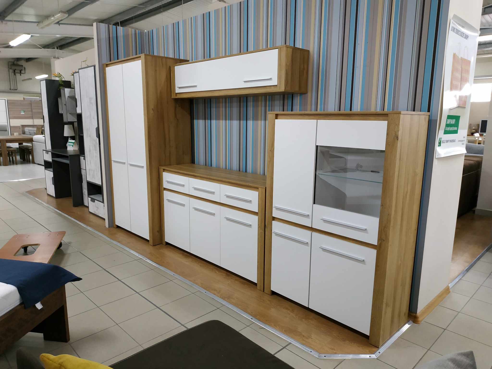 Komplet mebli pokojowych z jasnego drewna i białych frontów, składający się z szafy, szafek wiszących i stojących, prezentowany w salonie meblowym na tle ściany w pionowe pasy.