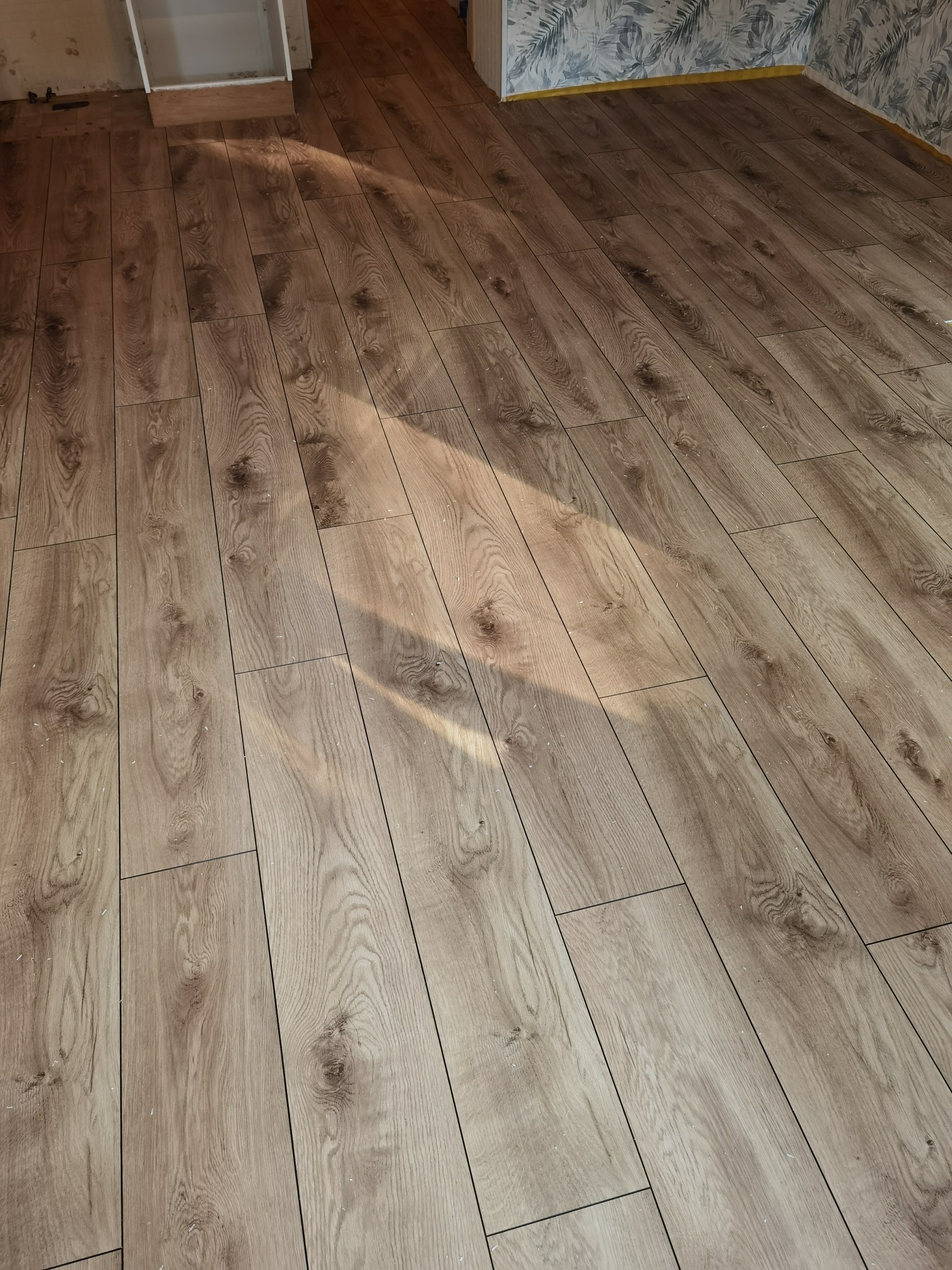 Ukończona podłoga z paneli laminowanych o wzorze jasnego dębu, ułożona wzdłuż, z widocznym fragmentem ściany z tapetą w liście w tle, oświetlona naturalnym światłem tworzącym smugę na powierzchni.