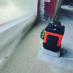 Laser krzyżowy emitujący zielone linie na szarej, szorstkiej wylewce podczas prac wykończeniowych, widoczne ślady po wierceniu i folia ochronna na ścianie.