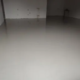 Świeżo wylana, gładka posadzka w jasnym odcieniu szarości, z widocznym niebieskim wężem na brzegu, schnąca w surowym wnętrzu z białymi ścianami.