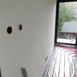 Wnętrze w trakcie prac wykończeniowych: widoczne czerwone rury ogrzewania podłogowego na folii, świeżo otynkowana ściana z otworami na gniazdka, poziomica przy ścianie, okno z widokiem na zewnątrz.