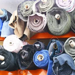 Magazyn w Łodzi: Rzędy różnorodnych tkanin zwiniętych w bele, ułożone na pomarańczowych półkach, widoczne faktury i kolory materiałów, od delikatnych pasteli po głębokie granaty i czernie.