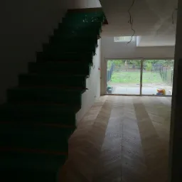 Wnętrze domu w trakcie remontu: zabezpieczone folią schody i parkiet ułożony w jodełkę, widoczne wiszące kable elektryczne.