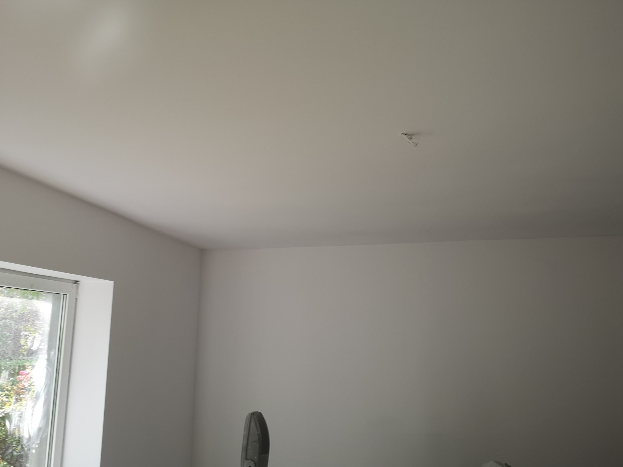 Ściana i sufit po malowaniu na jasny odcień szarości, widoczny fragment okna z roślinnością za szybą oraz wystający kabel elektryczny na suficie.