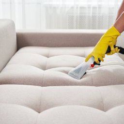 Fresh Factory - Pracownik w żółtej rękawicy czyści jasną, tapicerowaną kanapę za pomocą odkurzacza piorącego z przezroczystą końcówką, widoczne ślady wilgoci.
