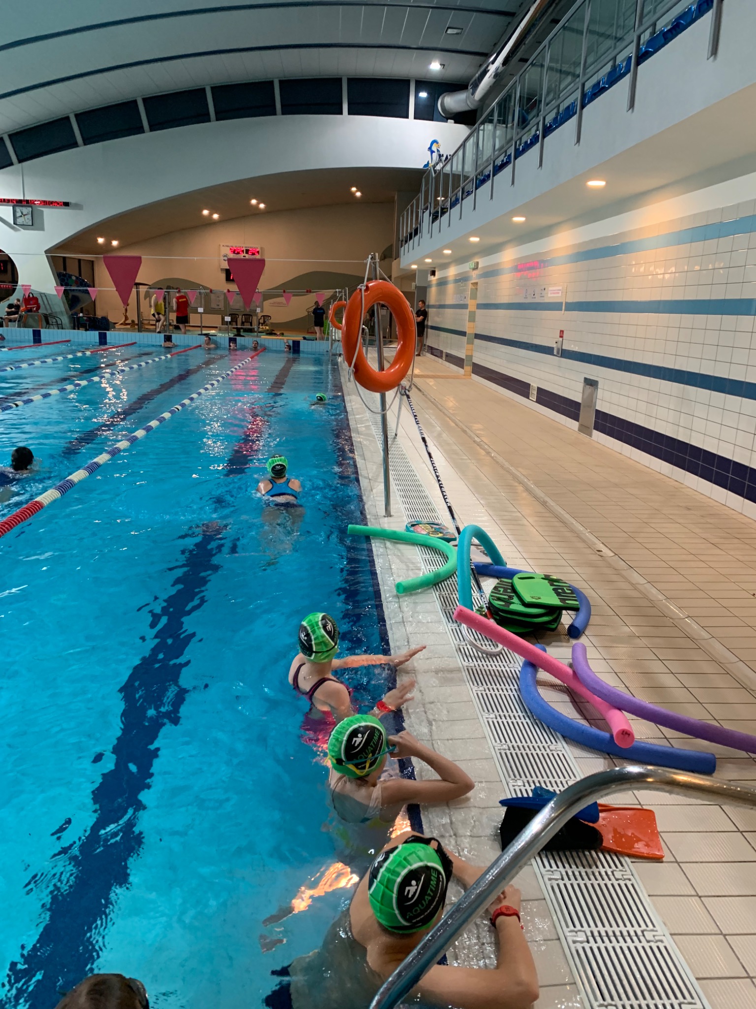 Kryty basen z torem pływackim, dzieci w zielonych czepkach z logo Aquatime przy krawędzi basenu, obok leżą kolorowe makarony do pływania i deski.